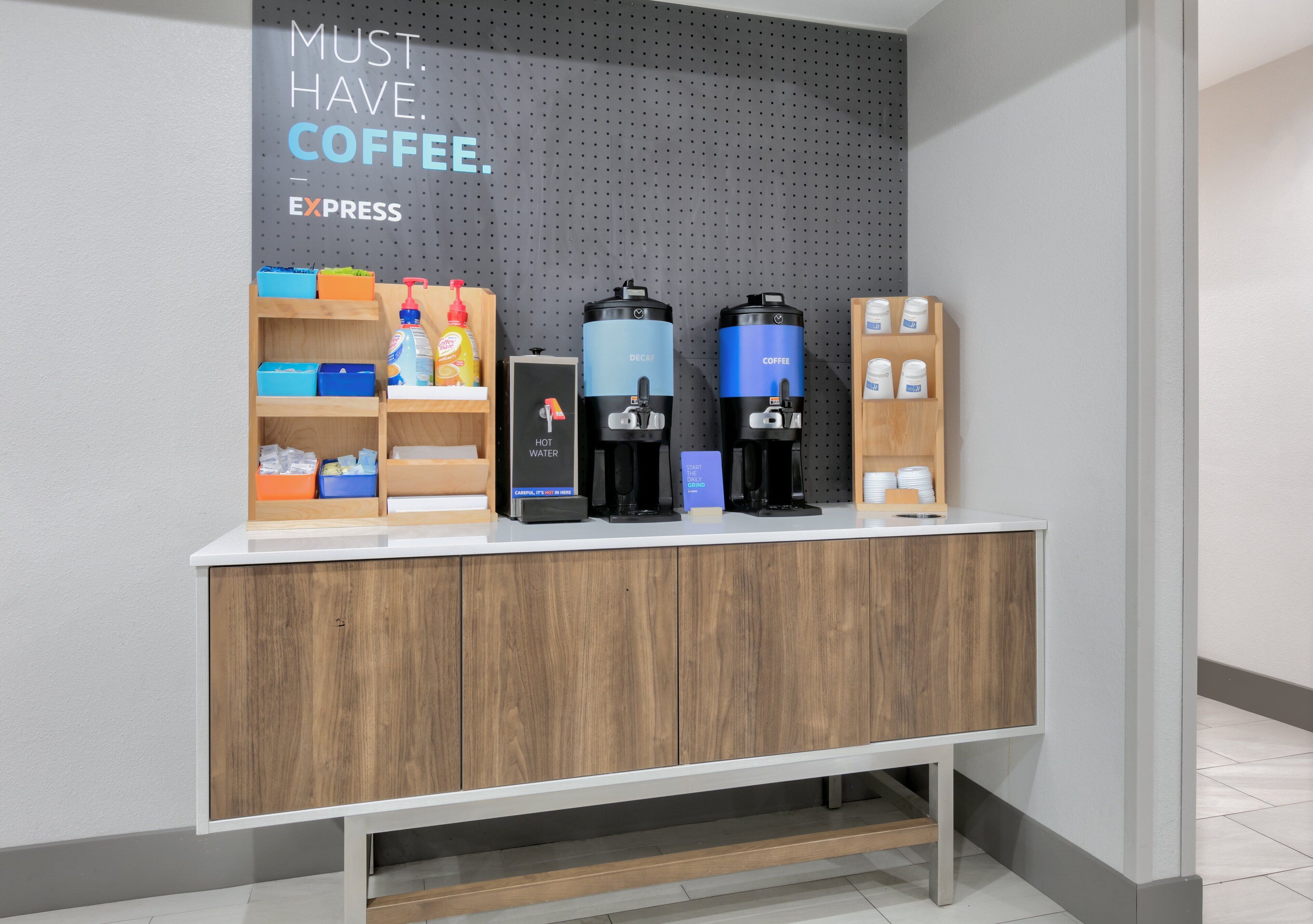A coffee station with dispensers labeled 'HOT WATER' and 'COFFEE' and a sign that reads 'MUST. HAVE. COFFEE. EXPRESS'.
