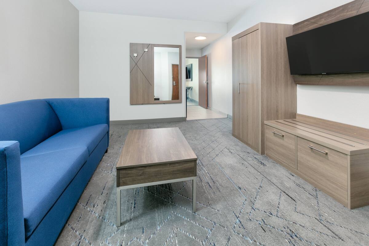A modern hotel room with a blue sofa, a wooden coffee table, a wardrobe, and a flat-screen TV mounted on the wall.