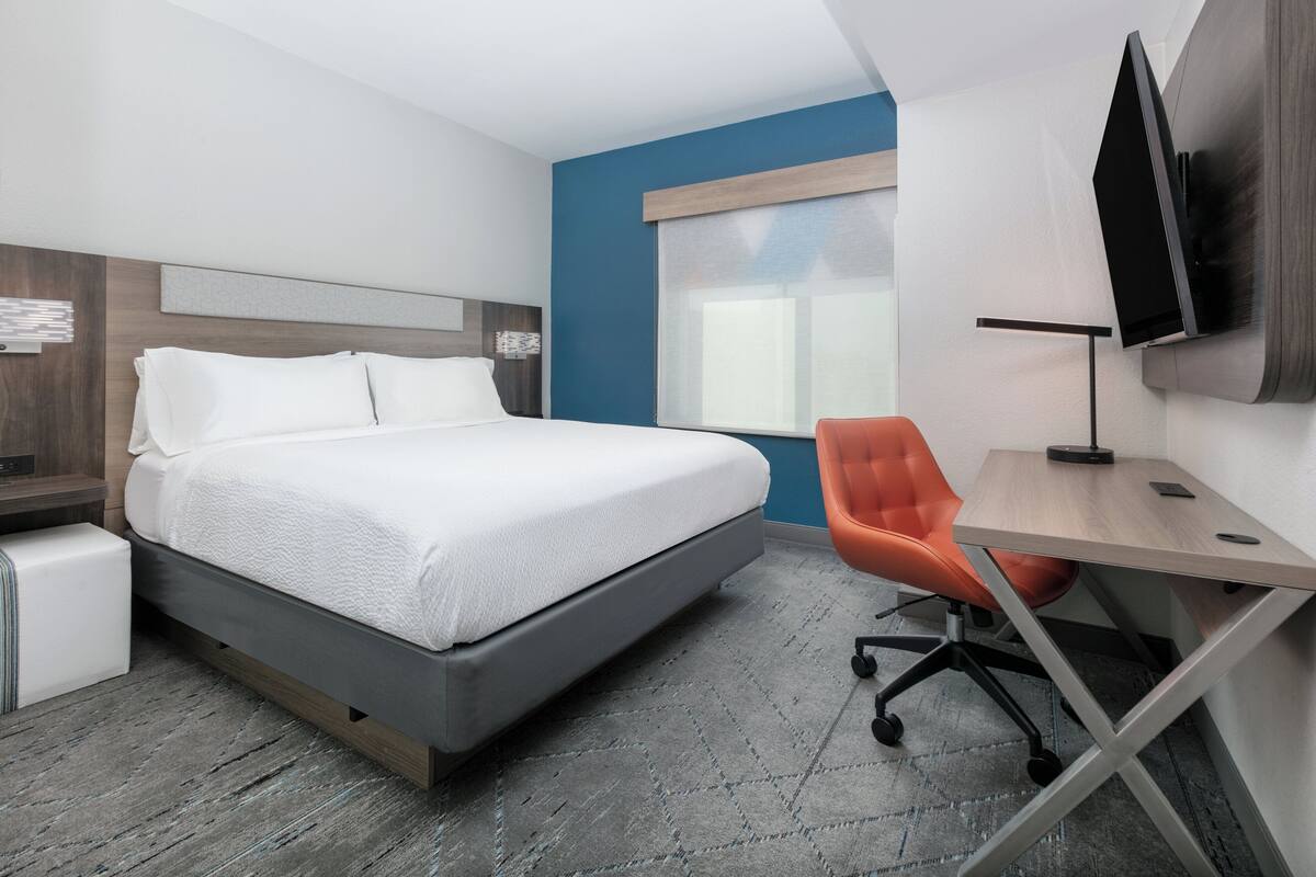 A modern hotel room with a bed, a desk, an orange chair, a TV, and a window.