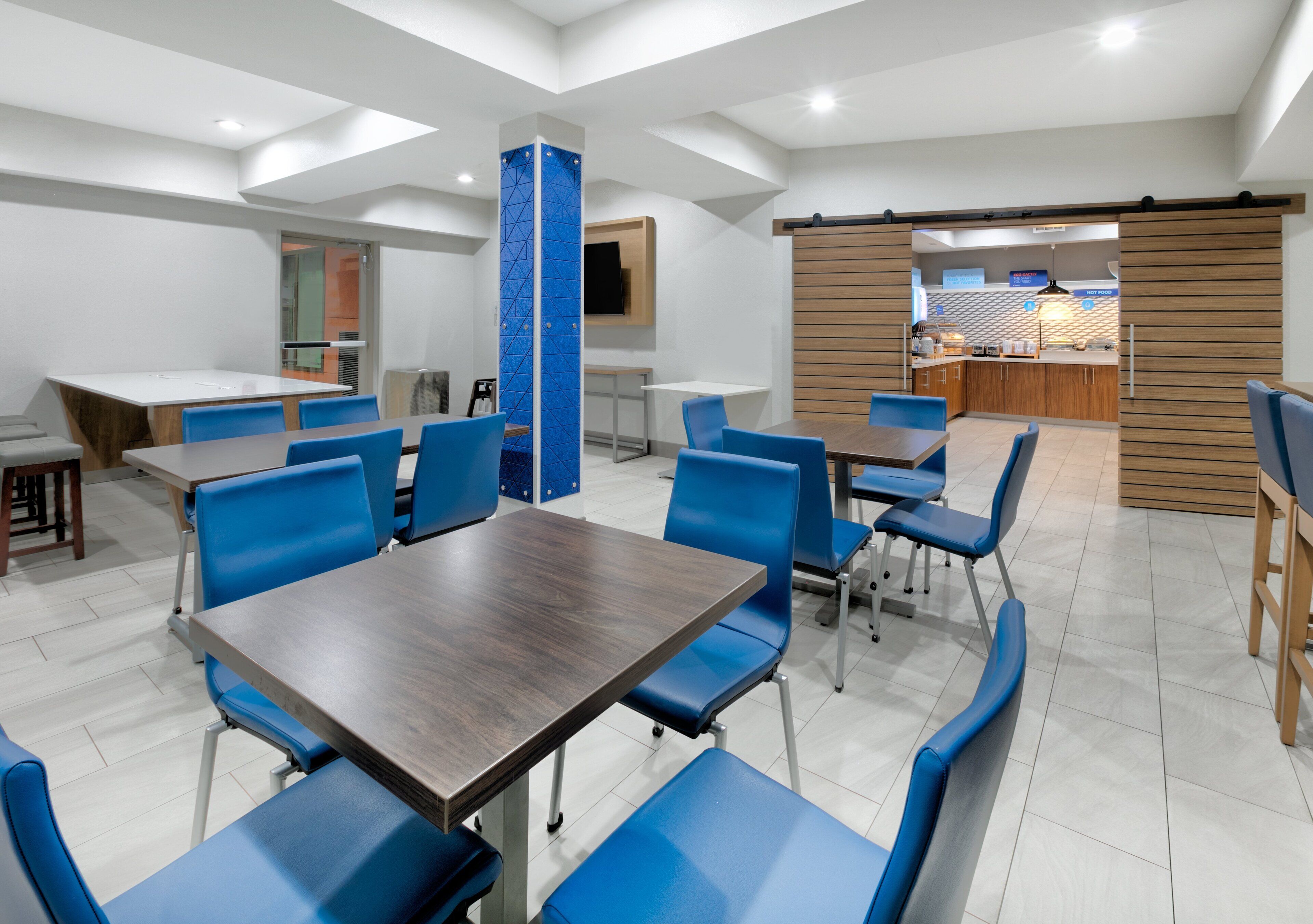 Un espace de restauration moderne dans un hôtel, avec de nombreuses tables et des chaises bleues, une cuisine en arrière-plan et une grande colonne ornée d’un motif géométrique.