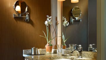 A modern bathroom with a granite countertop, a large mirror, two wall-mounted lights, a towel rack, and a potted orchid.