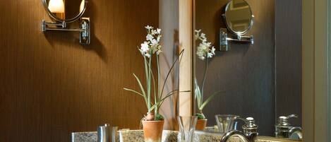 A modern bathroom with a granite countertop, a large mirror, two wall-mounted lights, a towel rack, and a potted orchid.