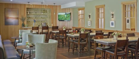 Un restaurante con varias mesas y sillas, una zona de bar con estantes llenos de vasos, y un televisor colgado en la pared.