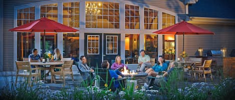 A group of people seated around a fire pit in an outdoor patio area with large windows and red umbrellas.