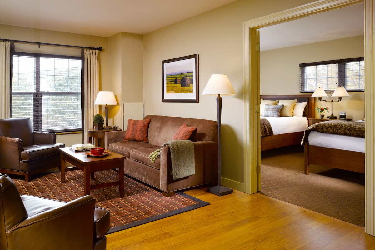 Ein Hotelzimmer mit braunem Ledersofa, einem Holztisch, einem kleinen Teppich, einem Bett mit Holz Kopfteil, zwei Nachttischlampen und einem Fenster mit Jalousien.