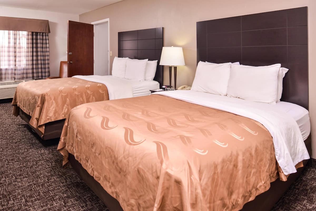 A hotel room with two beds, a nightstand, a lamp, and a window with curtains.