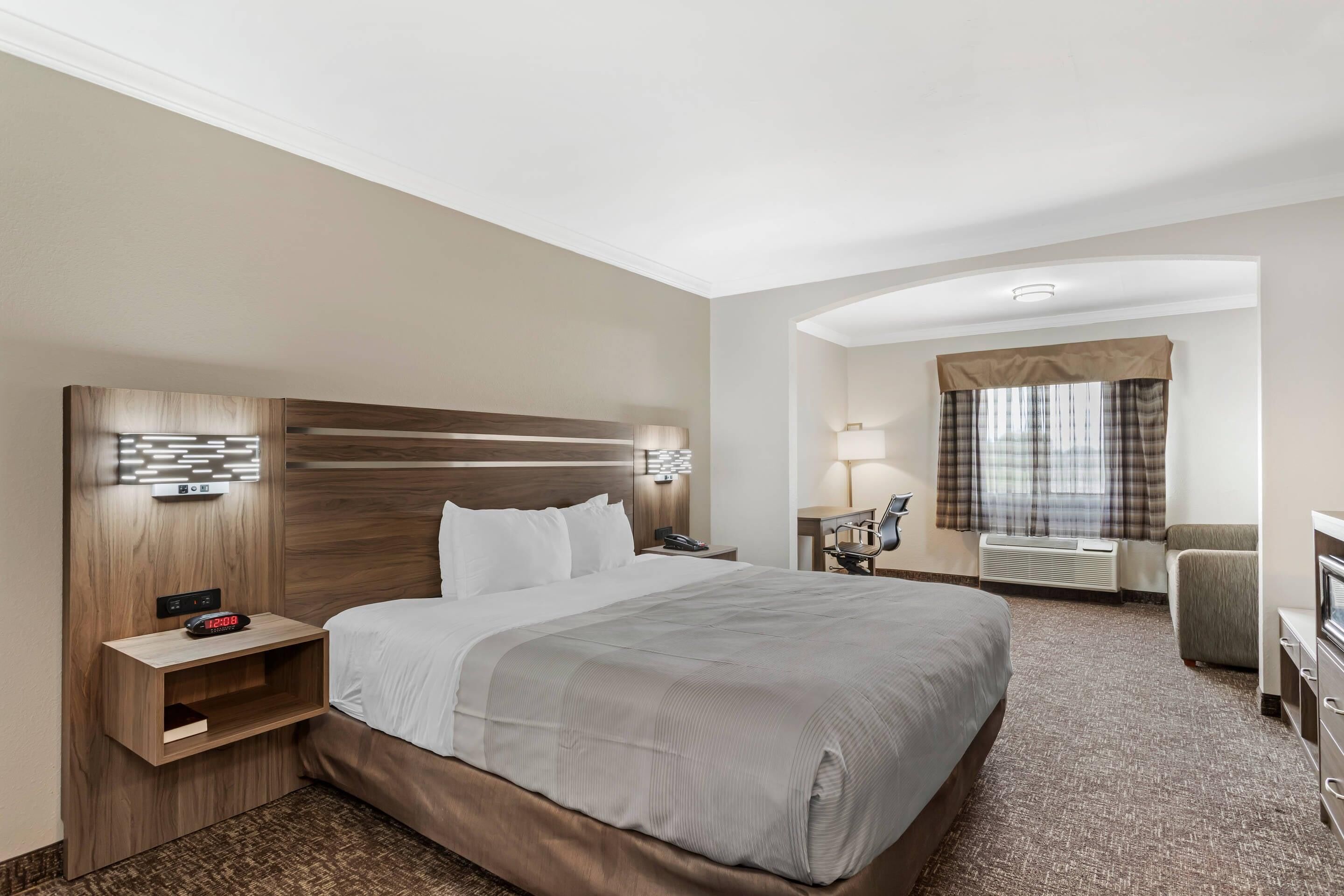 A hotel room with a large bed, a desk, a chair, a nightstand, and a wall-mounted lamp.
