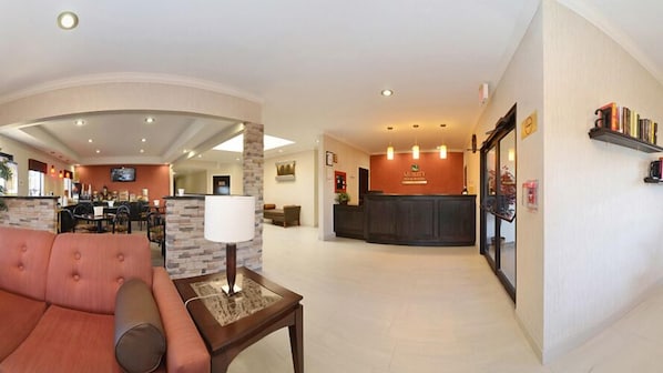Un hall de hotel con una recepción, una zona de estar con sofá y mesa, y un comedor al fondo.