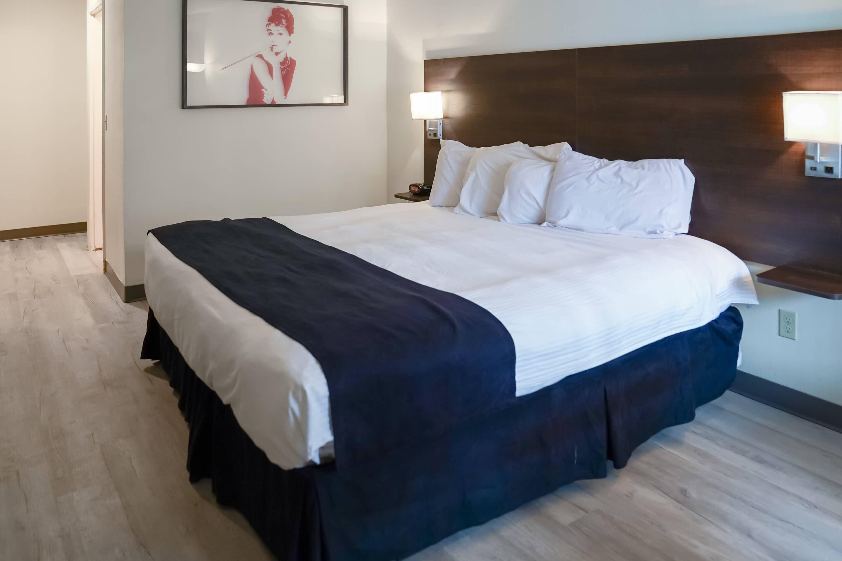 A hotel room with a large bed, a headboard, two bedside lamps, a framed picture on the wall, and a wooden floor.