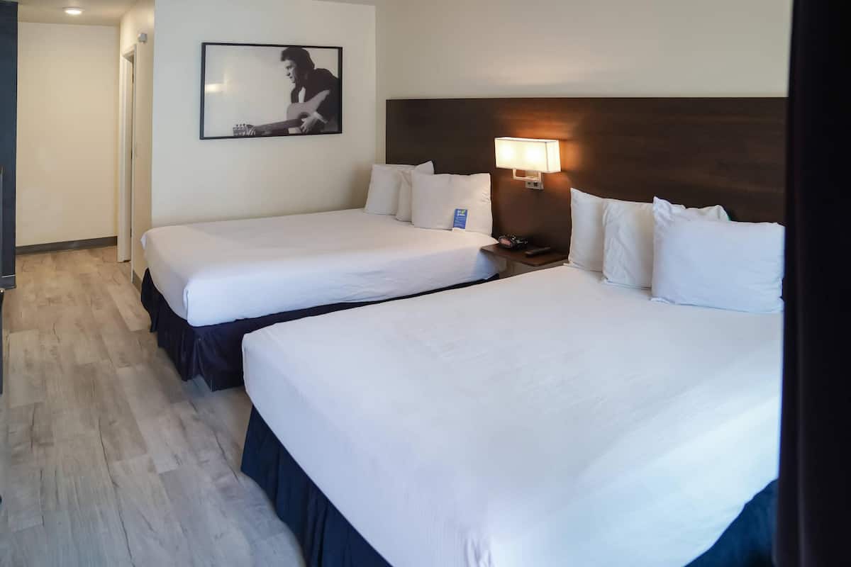 A hotel room with two beds, a nightstand with a lamp, a framed picture on the wall, and a wooden floor.