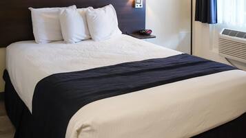 A hotel room with a bed, white pillows, a dark headboard, a bedside table with a lamp, and an air conditioning unit.