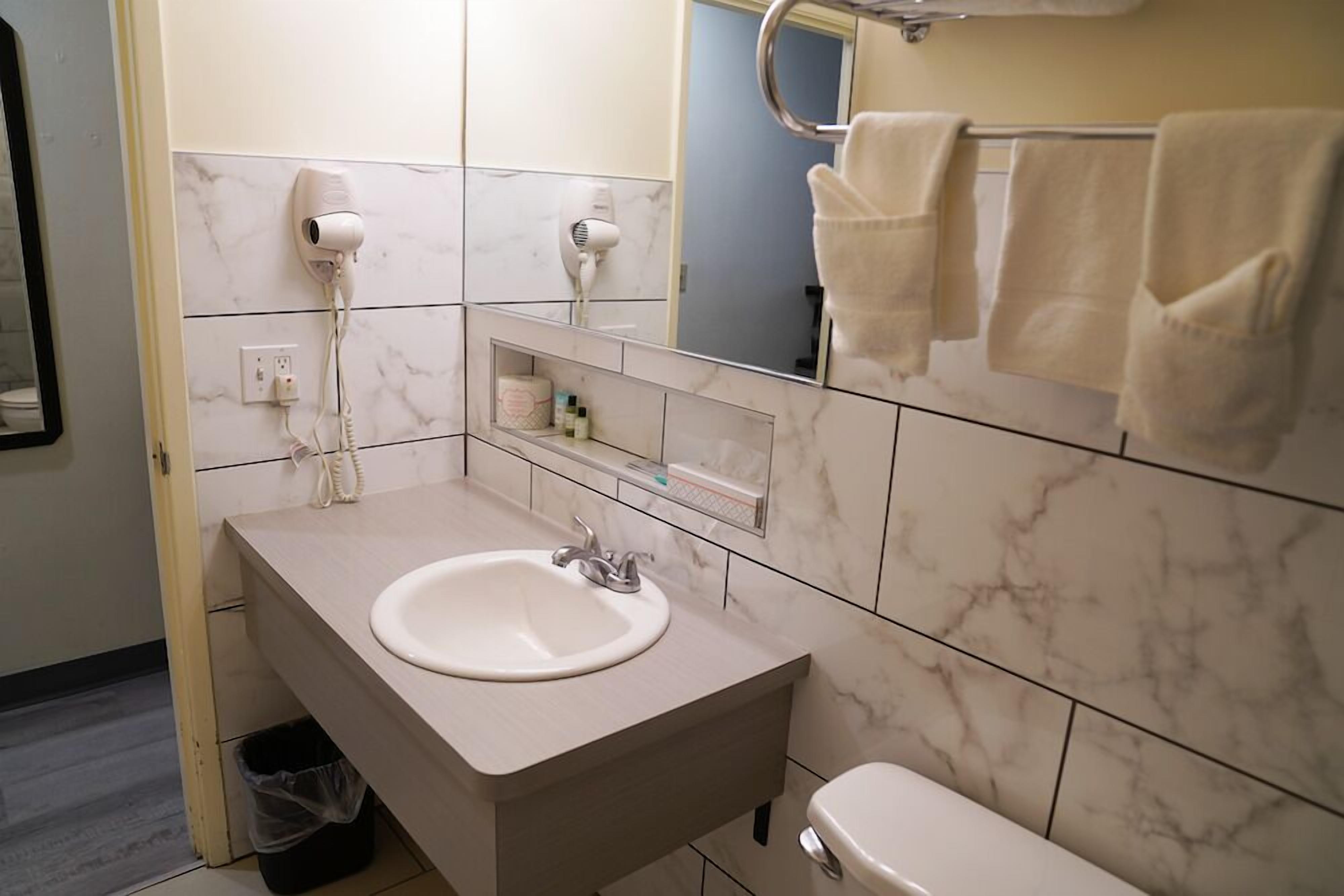 Ein Badezimmer mit Marmorfliesen, einem Waschbecken mit Spiegel darüber, zwei an der Wand befestigten Haartrocknern, einem Handtuchhalter mit Handtüchern und einem Mülleimer.