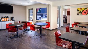 A modern dining area with red chairs and black tables, a kitchen area with a flat-screen TV, and a view of parked cars outside.