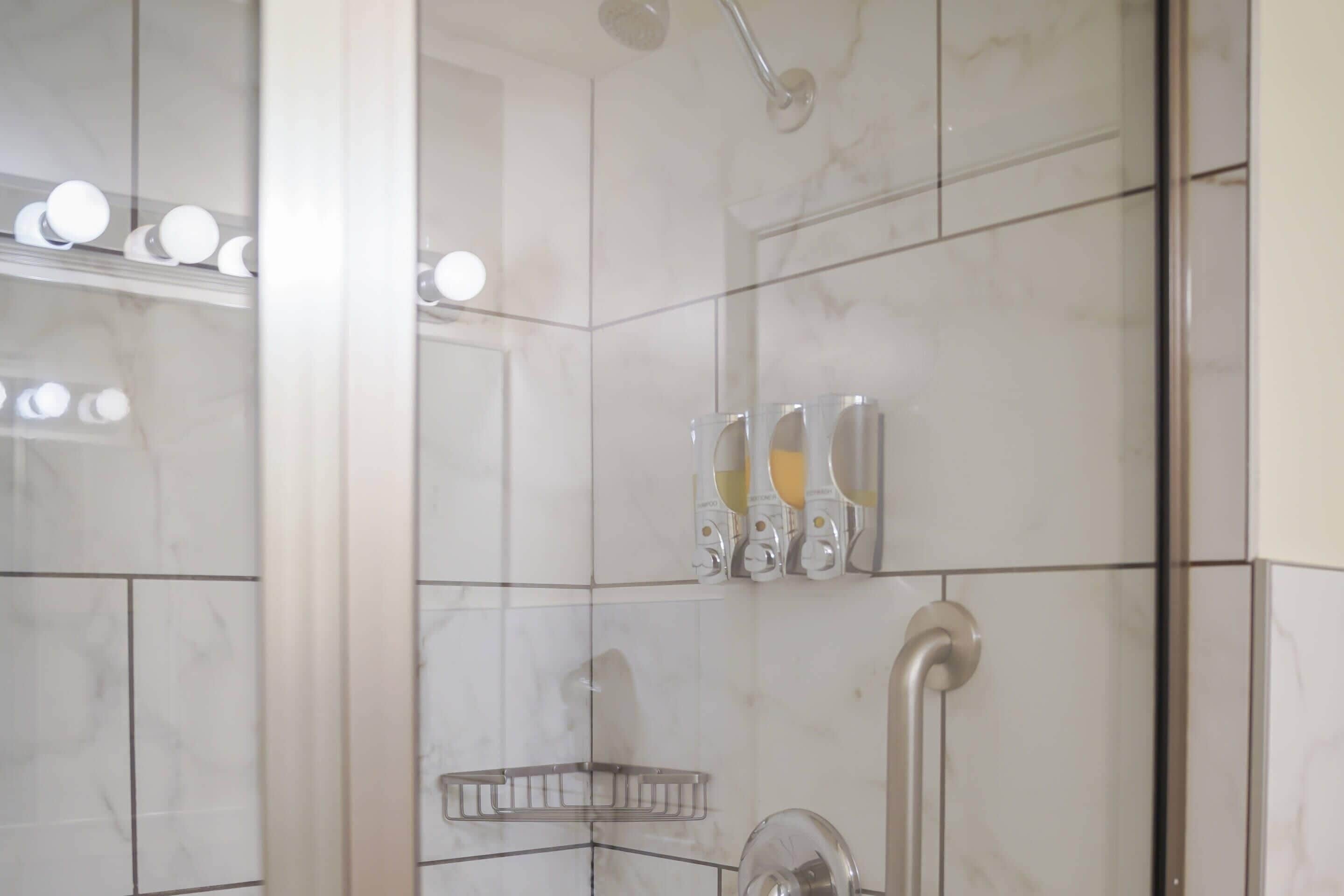 A bathroom with white marble tiles, a shower with a handheld showerhead, and a glass-enclosed mirror.