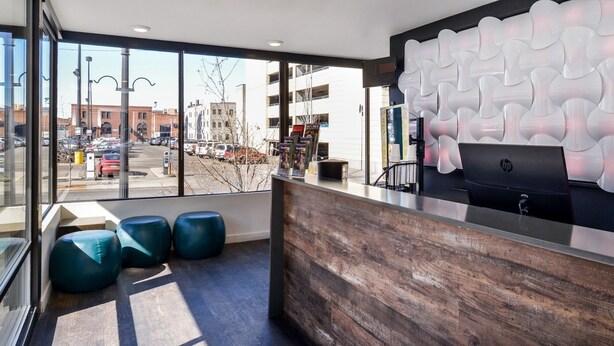 Un hall d’hôtel moderne avec un comptoir d’accueil en bois, une grande fenêtre donnant sur la rue et trois coins salons bleus avec des coussins.