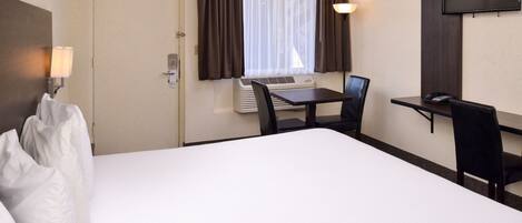 A hotel room with a bed, a desk, a chair, a television, and an air conditioning unit.