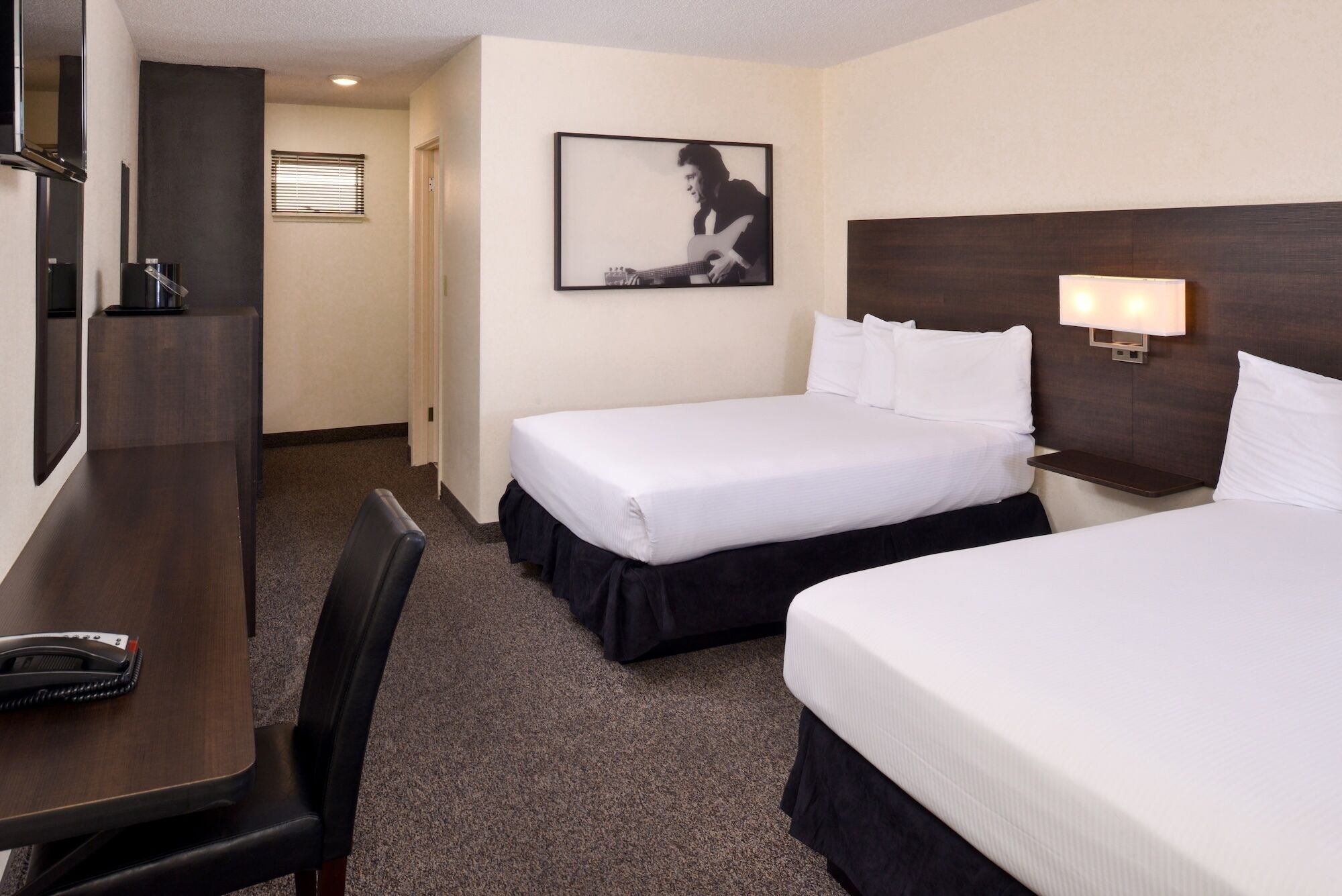 A hotel room with two beds, a desk, a chair, a framed picture on the wall, and a nightstand with a lamp.