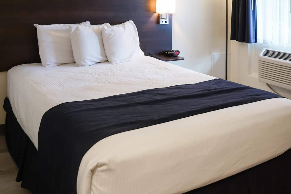 A neatly made bed with white and navy bedding, a bedside table with a lamp, and a wall-mounted air conditioner.
