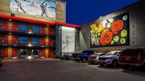 Un hôtel à étages, avec une fresque représentant des danseurs au deuxième étage et une grande fresque florale au troisième étage. Plusieurs voitures sont garées devant.