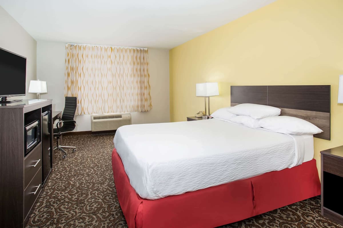 Ein Hotelzimmer mit einem großen Bett, einem Schreibtisch mit Fernseher, einem Sessel und einem Fenster mit Vorhängen.