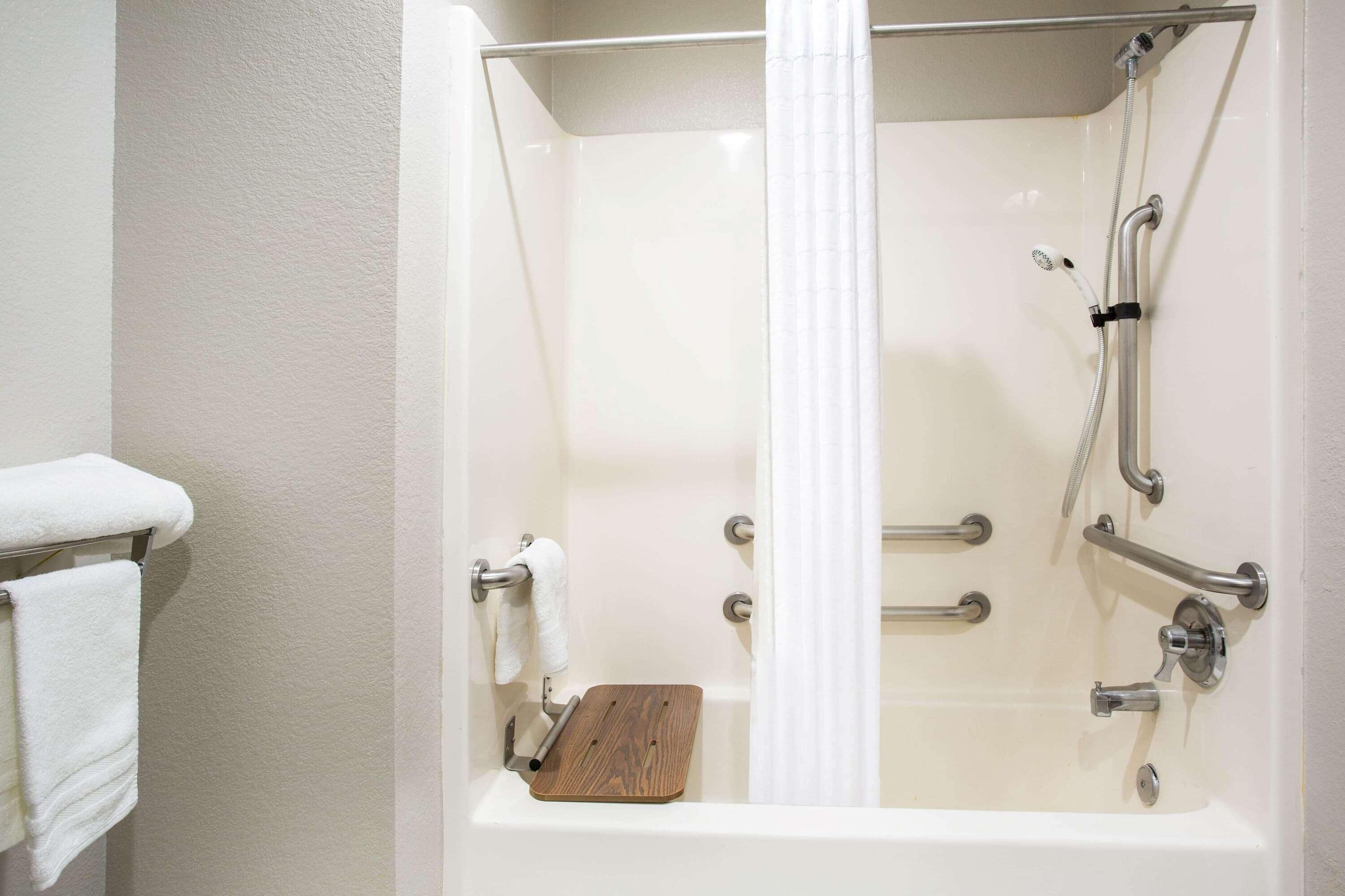 A bathtub with a wooden seat, grab bars, and a showerhead.