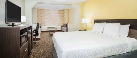 A hotel room with a large bed, a desk with a flat-screen TV, a chair, a sofa, and a small table.
