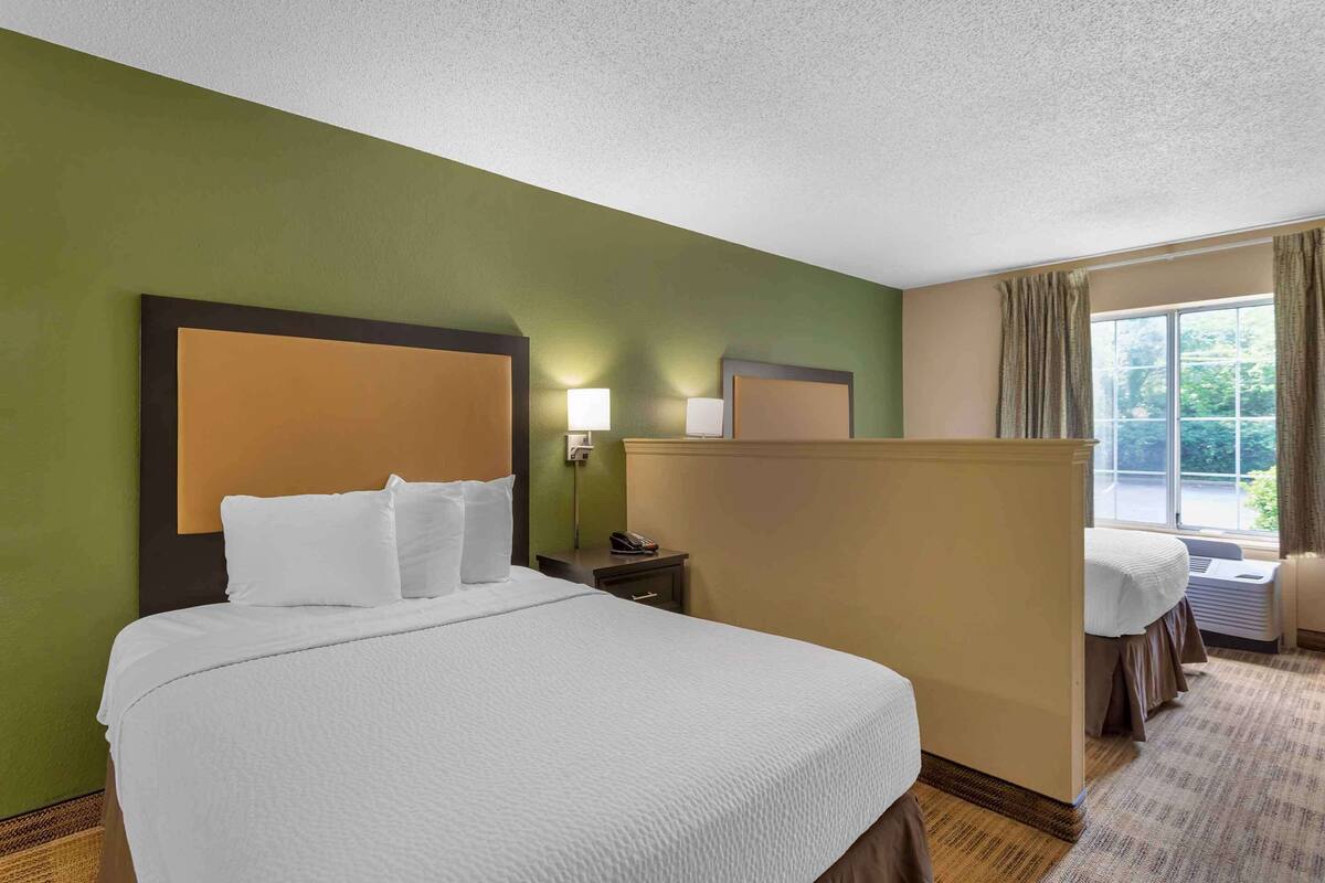 A hotel room with a large bed, a nightstand with a lamp, a window with curtains, and a view of greenery outside.