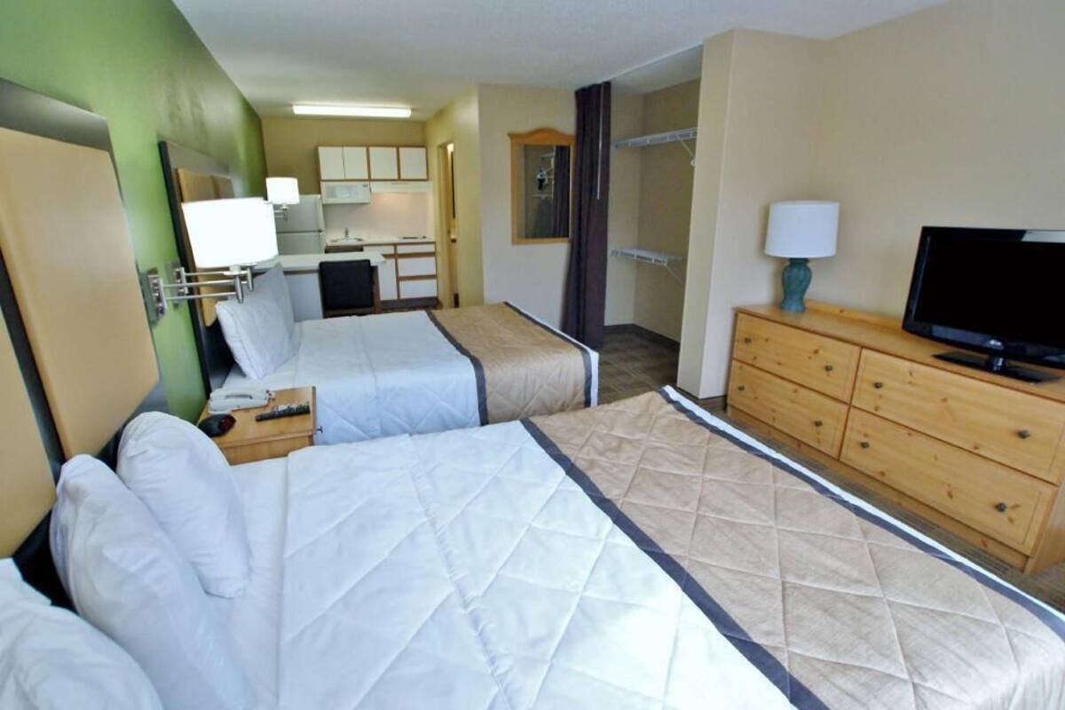A hotel room with two beds, a TV, a wooden dresser, and a kitchenette.