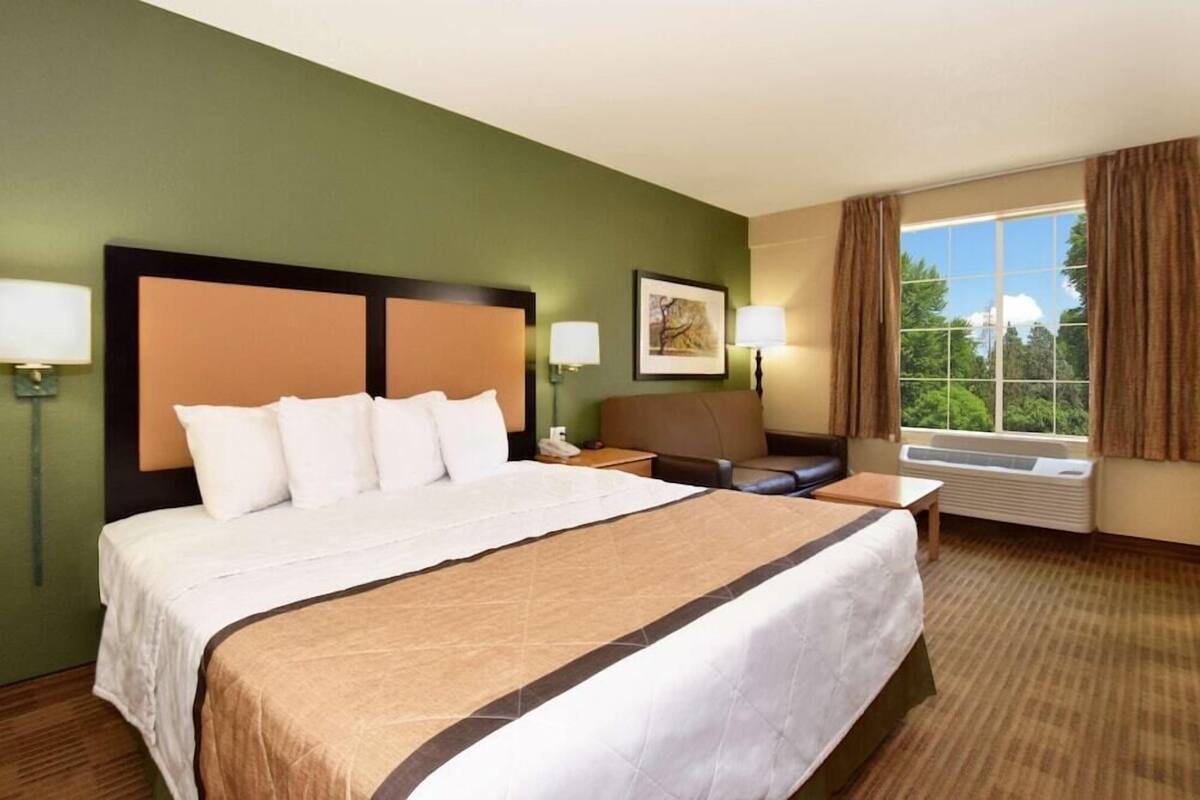 A hotel room with a large bed, a desk, a chair, a window with curtains, and a wall-mounted air conditioner.