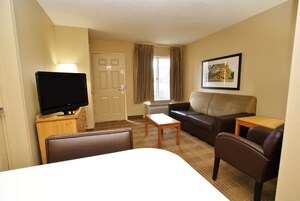 Una habitación de hotel con cama, televisor sobre un soporte de madera, sofá, pequeña mesa y ventana con cortinas.