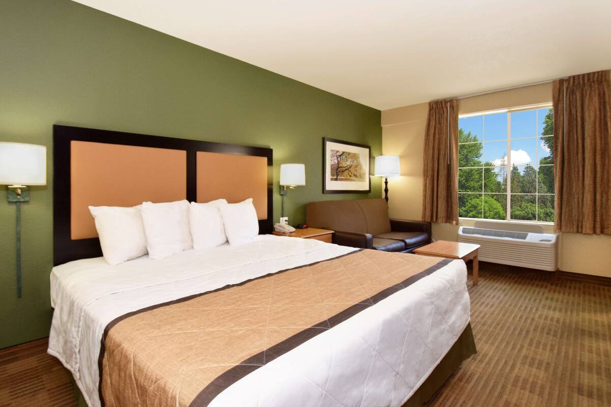 A hotel room with a large bed, a desk, a chair, a window with curtains, and a picture on the wall.