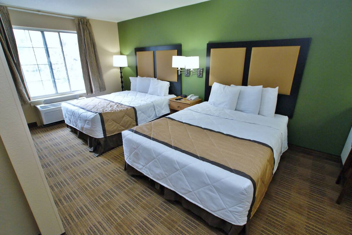 A hotel room with two beds, a window with curtains, a lamp, and a wall-mounted phone.