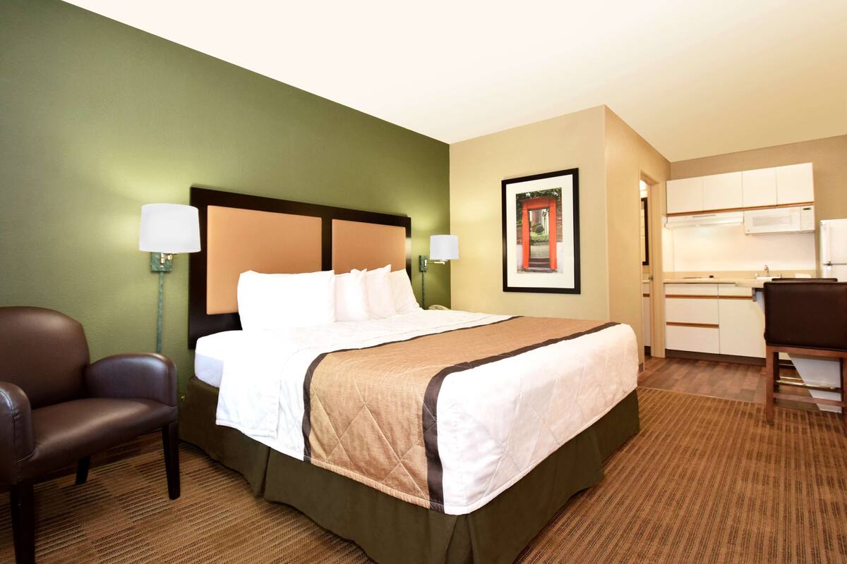A hotel room with a bed, a chair, a desk, and a kitchenette.