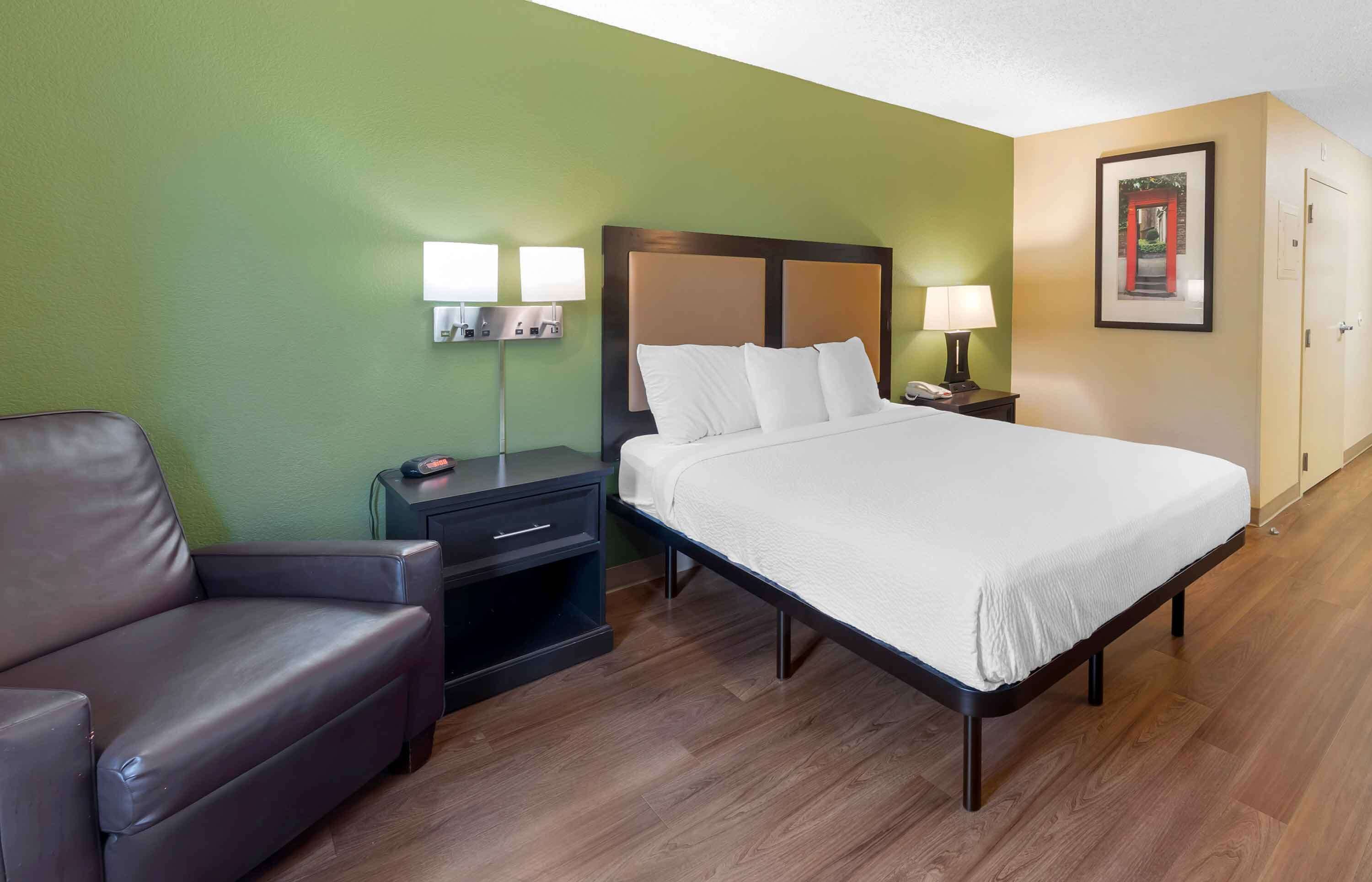 A hotel room with a bed, a nightstand, a lamp, a chair, and a framed picture on the wall.