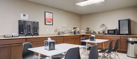 A kitchenette with a microwave, toaster, and coffee maker. There are four tables with chairs, and a trash can is visible.