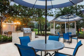 Een terras buiten met een ronde tafel en blauwe stoelen, een barbecue en een prieeltje.