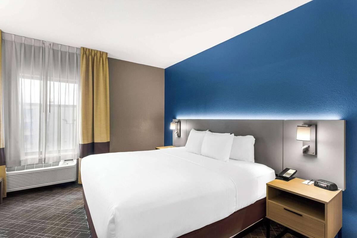 Ein Hotelzimmer mit einem großen Bett, einem Nachttisch mit Telefon, einer Wandleuchte und einem Fenster mit transparenten Vorhängen.
