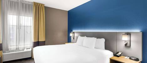 A hotel room with a large bed, a bedside table with a phone, a wall-mounted light, and a window with sheer curtains.