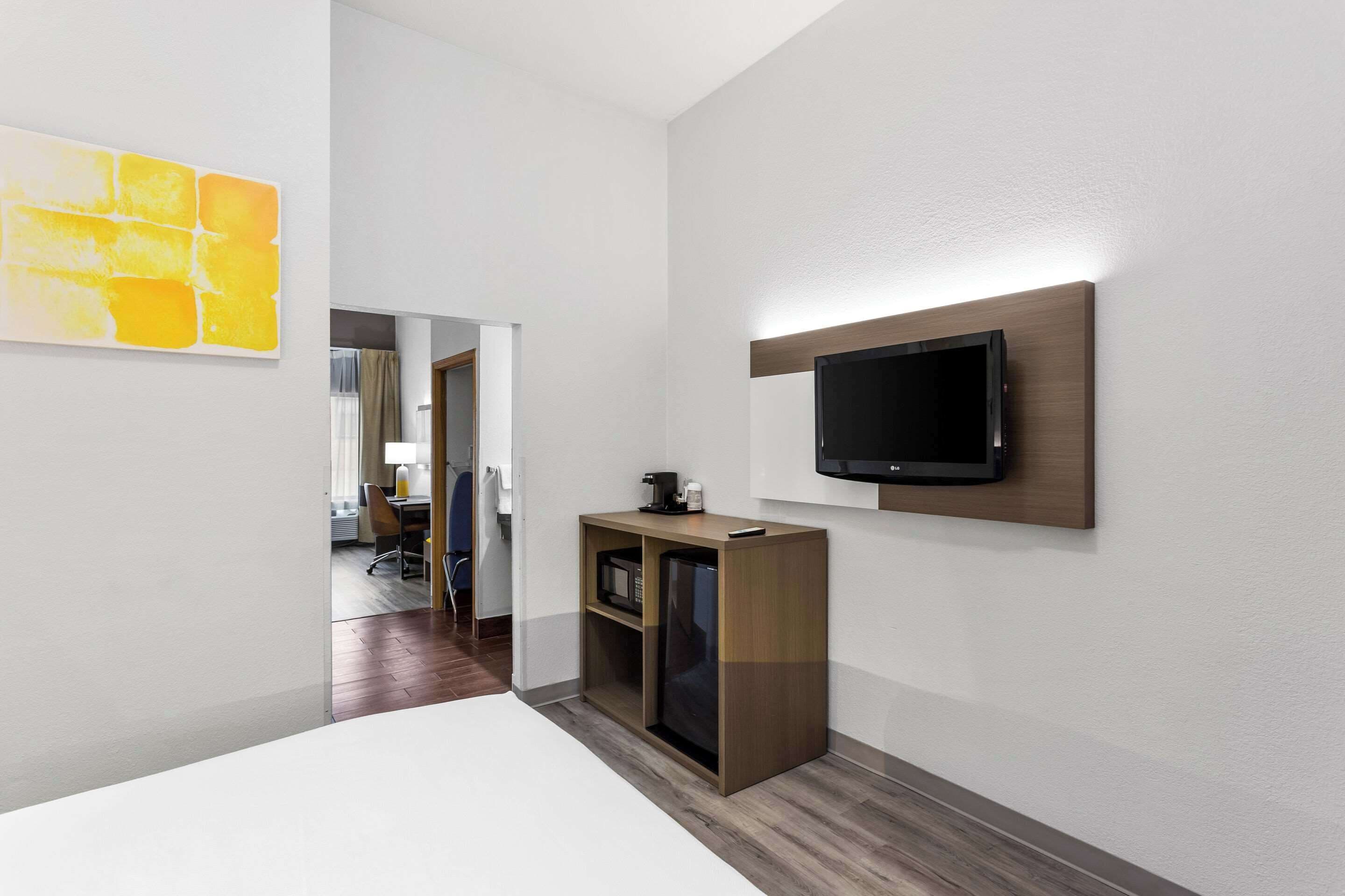 A modern hotel room with a bed, a TV mounted on the wall, a small wooden cabinet, and a bright yellow artwork on the wall.