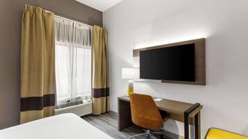 Habitación de hotel con cama, escritorio con silla, televisor de pantalla plana, lámpara y ventana con cortinas.