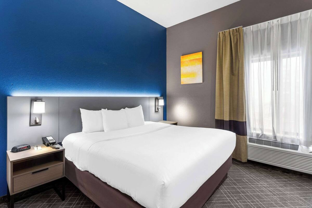 Ein Hotelzimmer mit einem großen Bett, Nachttischen mit Lampen, einem Wandtelefon und einem Fenster mit durchsichtigen Vorhängen.