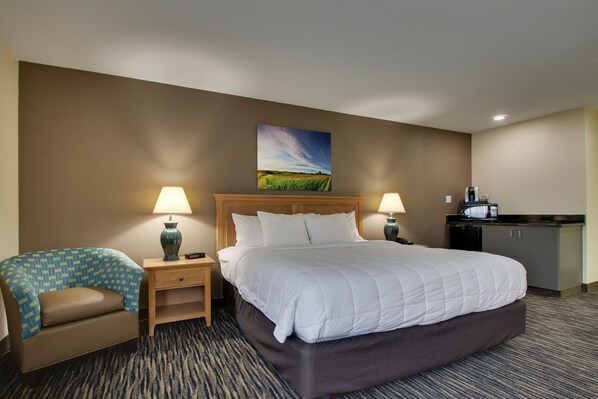 A hotel room with a large bed, two bedside tables with lamps, a small desk, a kitchenette, and a painting on the wall.