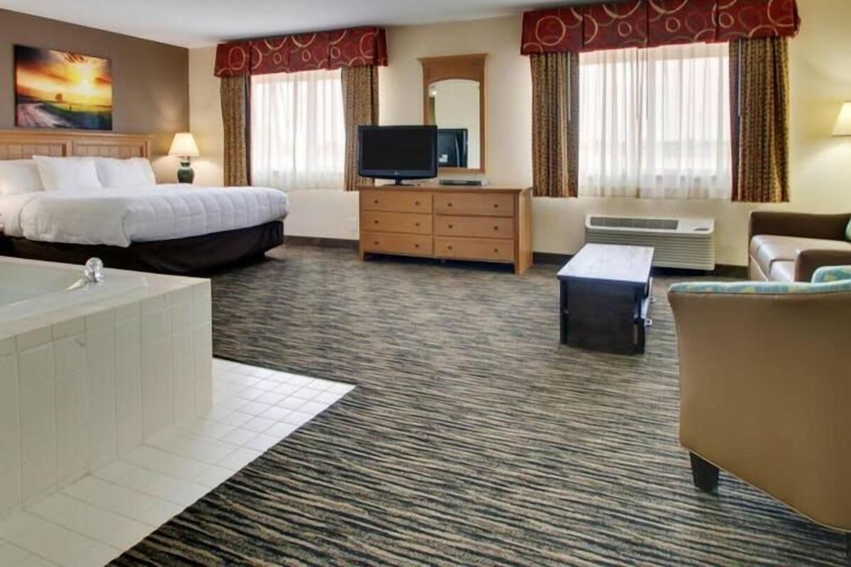Habitación de hotel con cama, escritorio, televisor, silla, sofá, baño con bañera y ventana con cortinas.