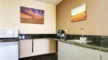 A compact kitchen with a microwave, coffee maker, sink, and a refrigerator. There are two paintings on the wall depicting landscapes.