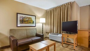 A hotel room with a brown leather sofa, a wooden coffee table, a flat-screen TV on a wooden stand, a framed picture on the wall, and a window with curtains. - Southpoint Suites Jacksonville (Jacksonville)