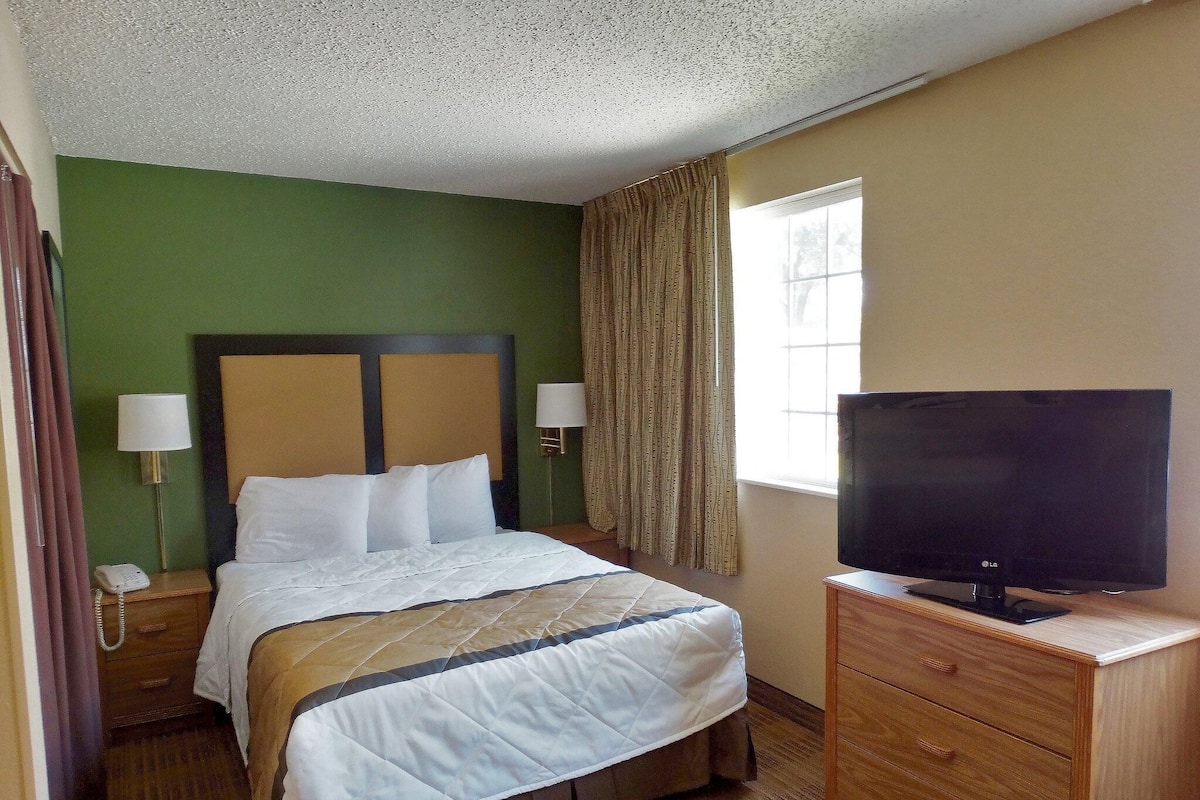 A hotel room with a bed, bedside lamps, a TV on a wooden dresser, and a window with curtains.