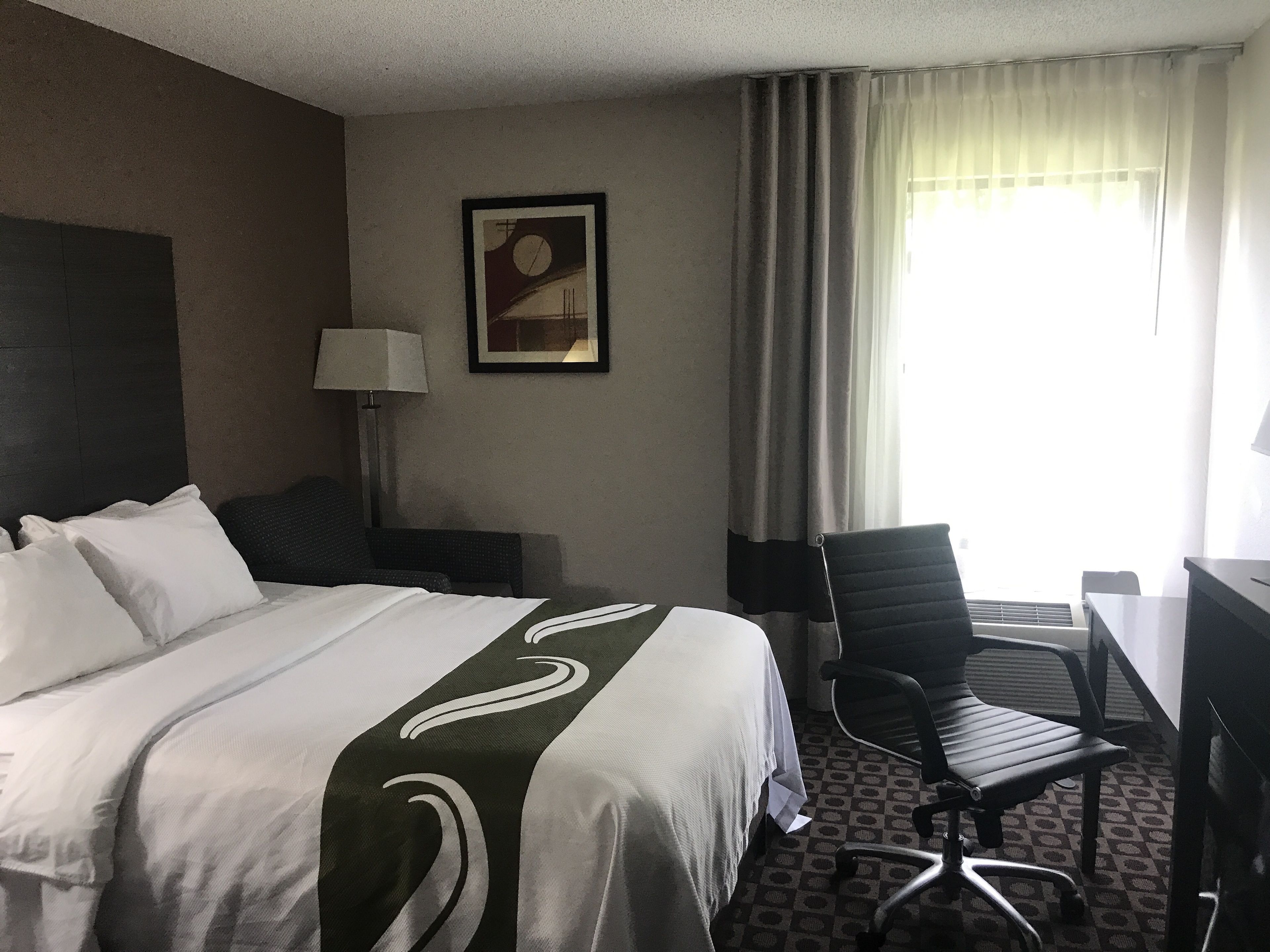 A hotel room with a bed, a desk, a chair, a lamp, and a painting on the wall.