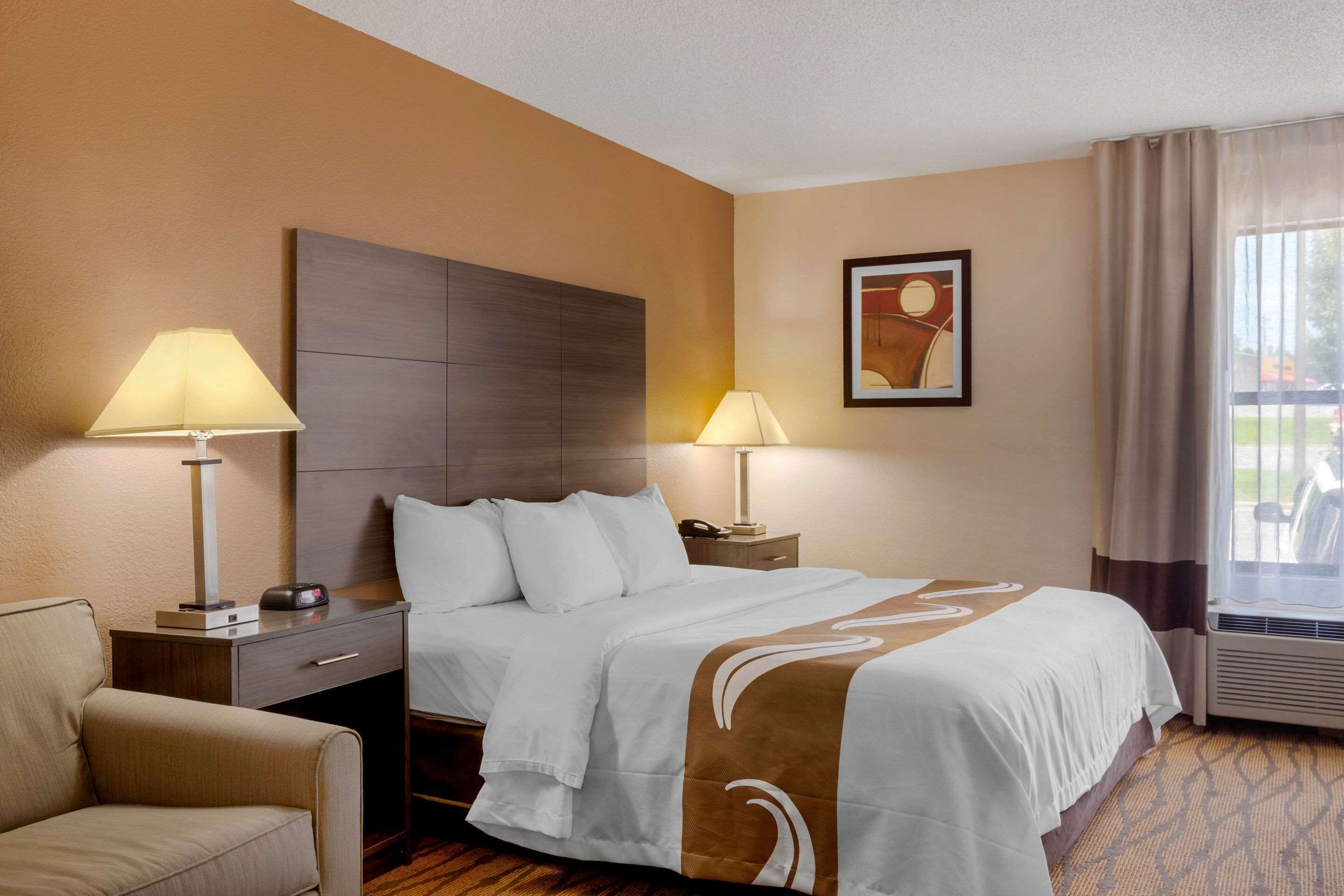 A hotel room with a bed, two bedside lamps, a nightstand, a chair, a framed picture on the wall, and a window with curtains.