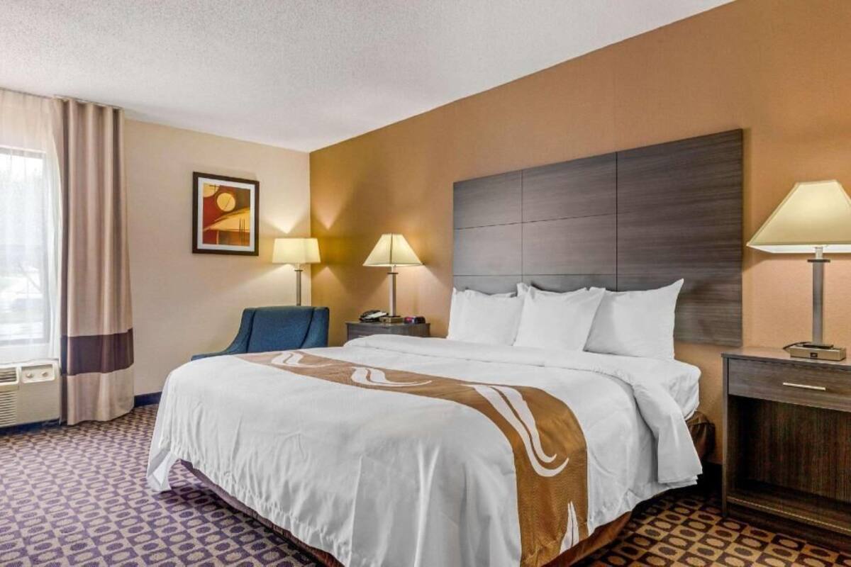 Hotellihuone, jossa on suuri sänky, kaksi yöpöytää, tuoli, työpöytä, lamppu ja taulu seinällä.