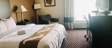 Habitación de hotel con una cama grande, un escritorio con computadora, una silla, una lámpara y un cuadro en la pared.